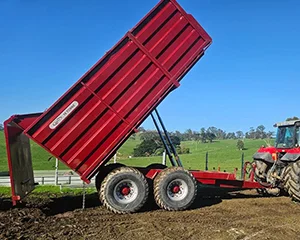 A red farm trailer tilted upward while unloading soil, attached to a tractor on a sunny day with green hills in the background.