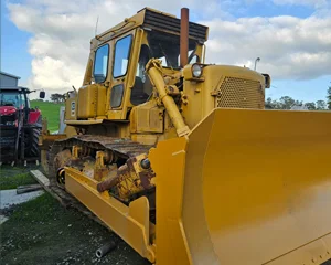 A large yellow bulldozer with a wide front blade parked outdoors on grass, with a red tractor visible in the background.