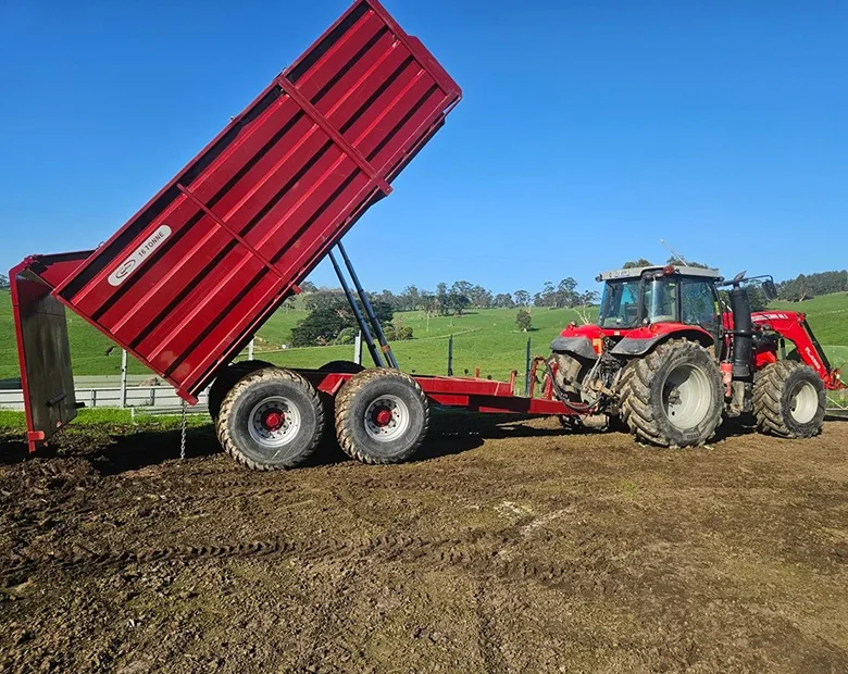 A red farm trailer tilted upward while unloading soil, attached to a tractor on a sunny day with green hills in the background.