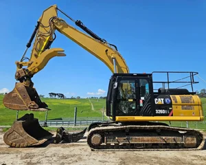 A large yellow CAT excavator parked on a dirt site under a clear blue sky, with green fields and trees in the background.