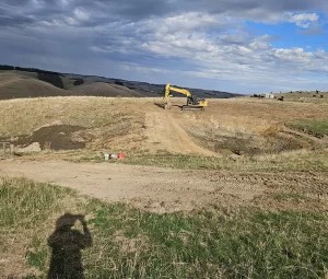 A yellow excavator works on a dirt excavation site in dry grassy hills under a partly cloudy sky, with a shadowed figure nearby.