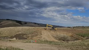 A yellow excavator works on a dirt hill under a cloudy sky, surrounded by dry grass and distant rolling farmland.