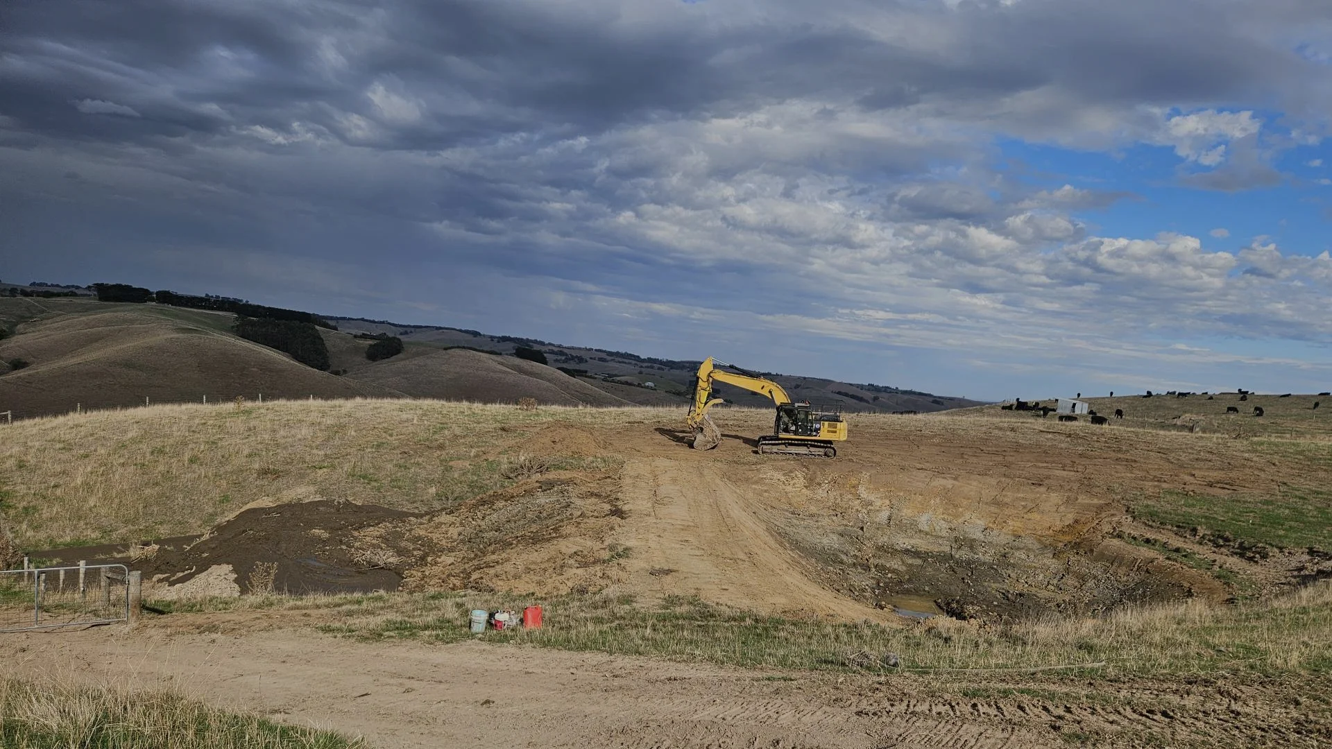 A yellow excavator works on a dirt hill under a cloudy sky, surrounded by dry grass and distant rolling farmland.